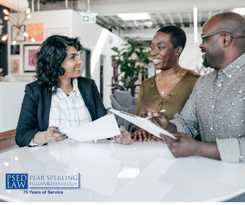 Three people in a modern office meeting around a white table, smiling and reviewing printed documents with one person pointing at a sheet.