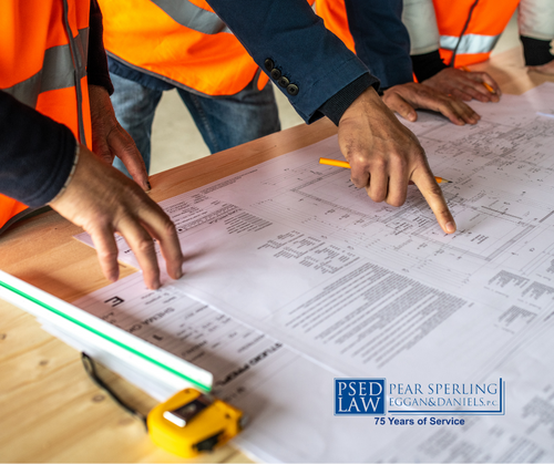 Construction workers in orange safety vests point at and review detailed blueprints spread on a wooden table, with hands tracing lines across the drawings.